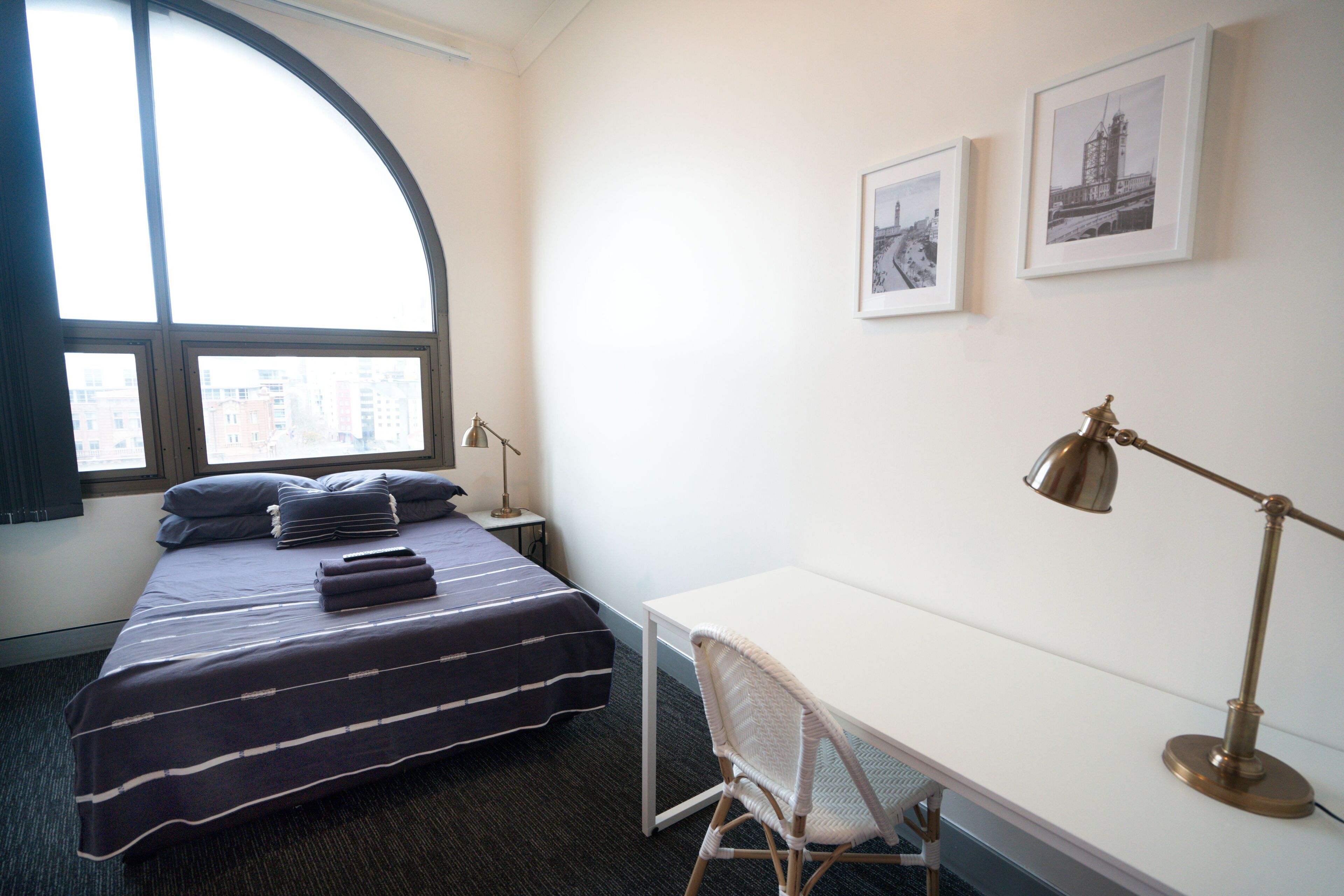 A hotel room with a bed, a desk, a chair, a lamp, and two framed pictures on the wall.