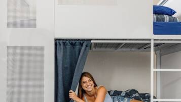 Un lit superposé avec une personne allongée sur le lit du bas, une valise et une fenêtre avec des rideaux.