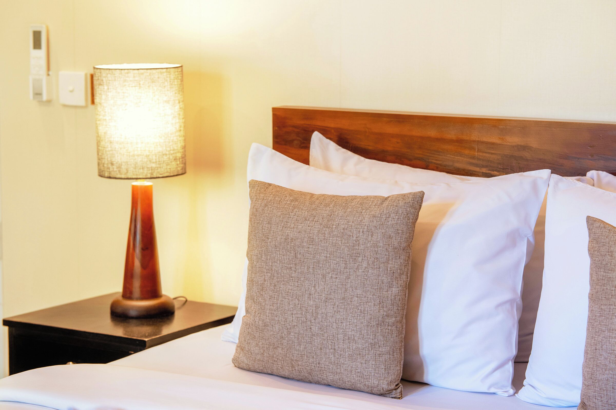 A neatly made bed with white and beige pillows, a wooden headboard, and a lamp on a bedside table.