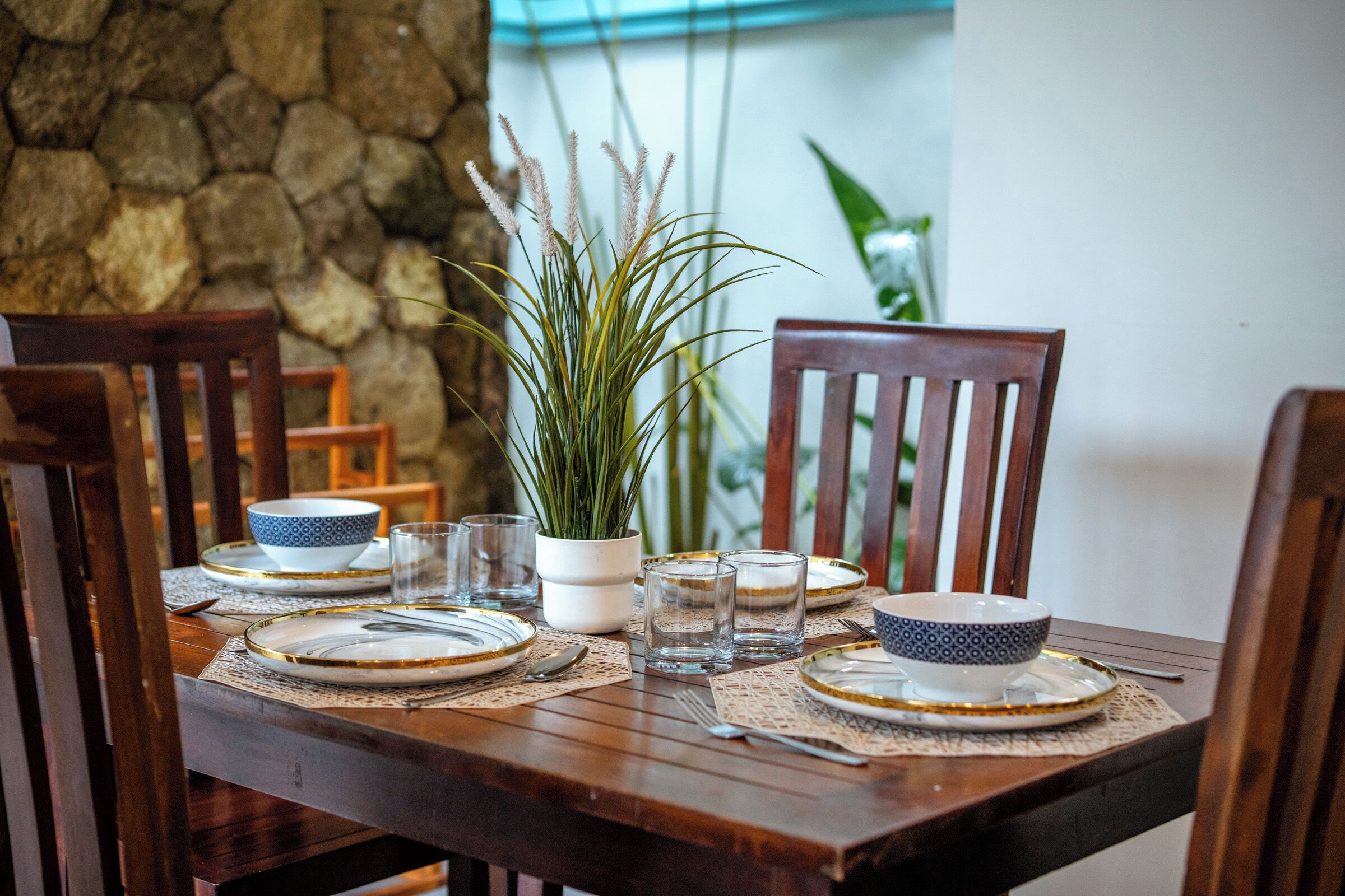 Una mesa de comedor de madera dispuesta con platos, vasos y una planta en maceta.