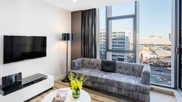 Een moderne woonkamer met een flatscreen televisie, een grijze bank met zwarte kussens, een salontafel met een tijdschrift en een vaas met gele bloemen, en een groot raam met uitzicht op de stad.