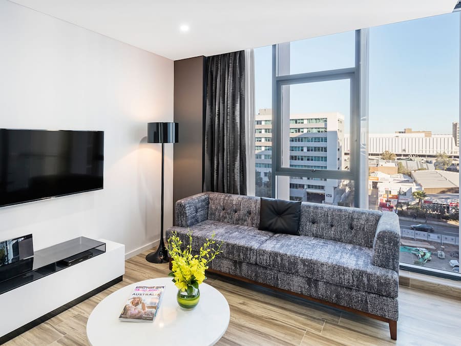 Ein modernes Wohnzimmer mit Fernseher, einem grauen Sofa mit schwarzen Kissen, einem Couchtisch mit einer Zeitschrift und einem Strauß gelber Blumen, und einem großen Fenster mit Blick auf die Stadt.
