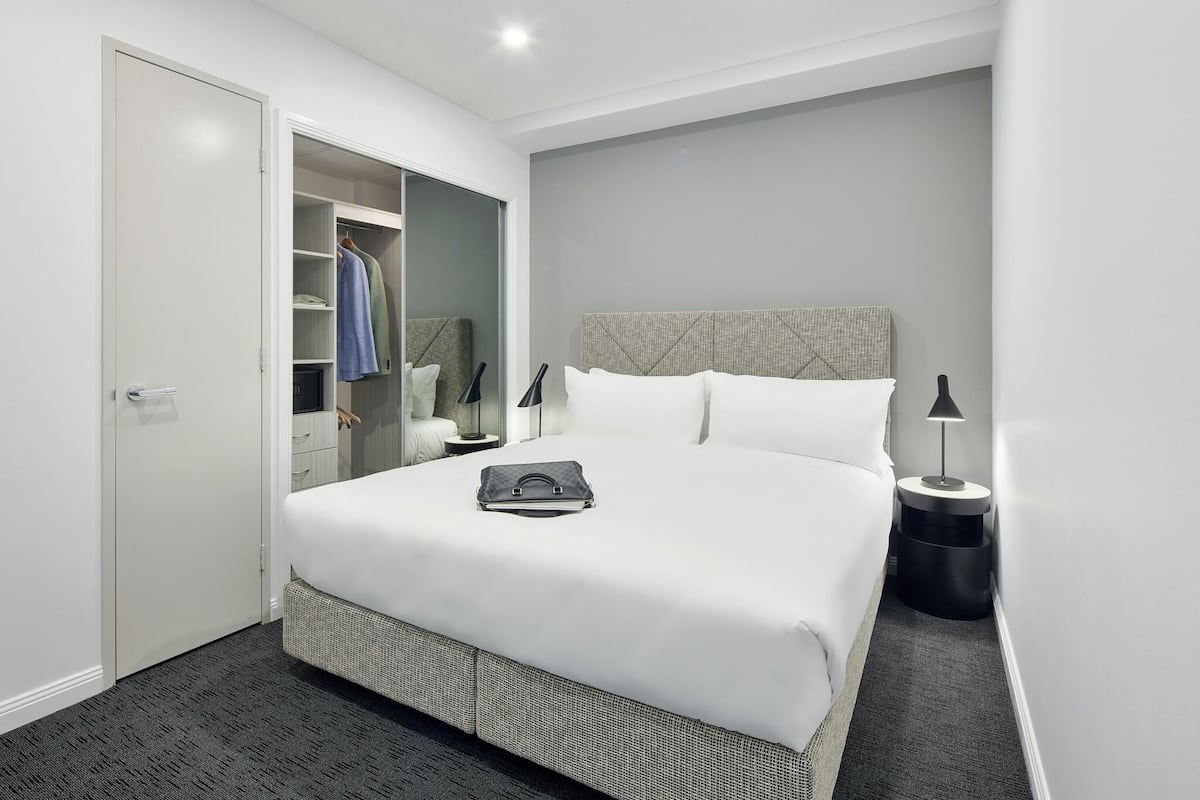 Una habitación de hotel moderna con una cama grande, un vestidor, una mesita de noche y una lámpara.