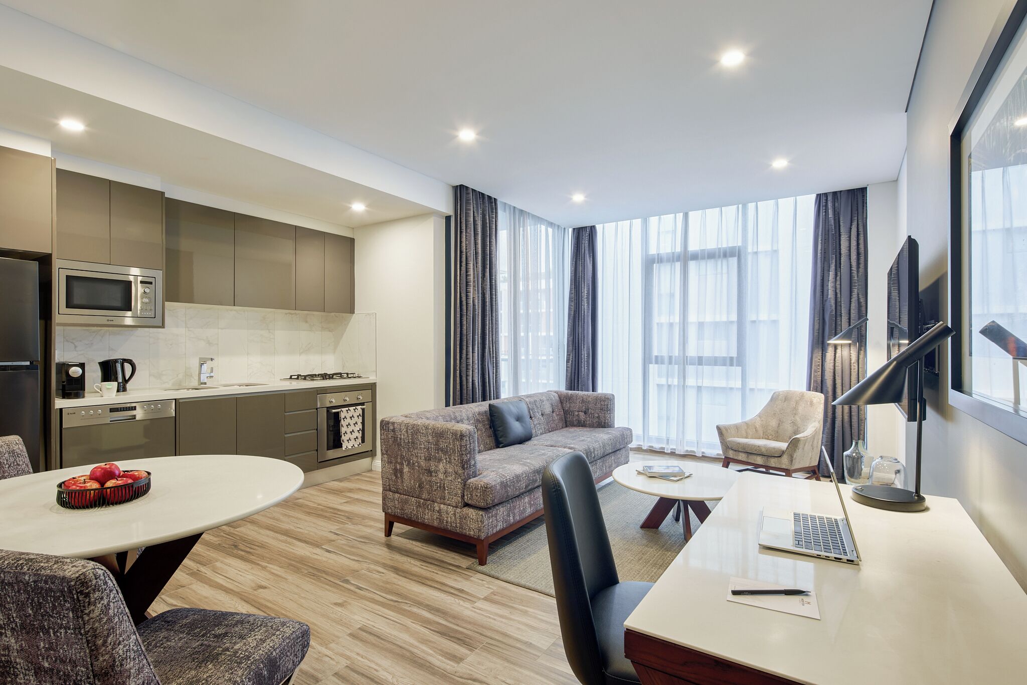 A modern hotel room with a kitchenette, a sofa, a dining table, and a desk with a laptop.
