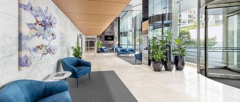 A modern hotel lobby with a marble wall, blue armchairs, potted plants, and a reception desk.