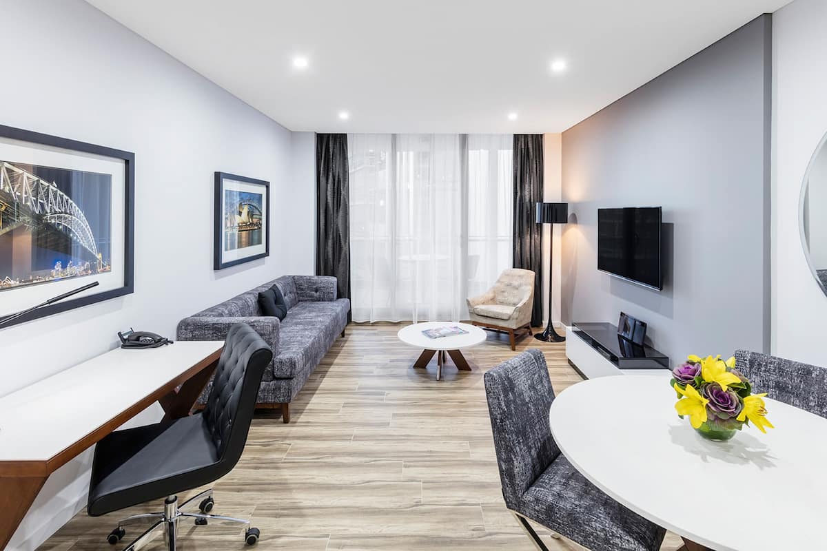Una habitación de hotel moderna con sofá, una mesa de comedor, un televisor y un amplio ventanal con cortinas.