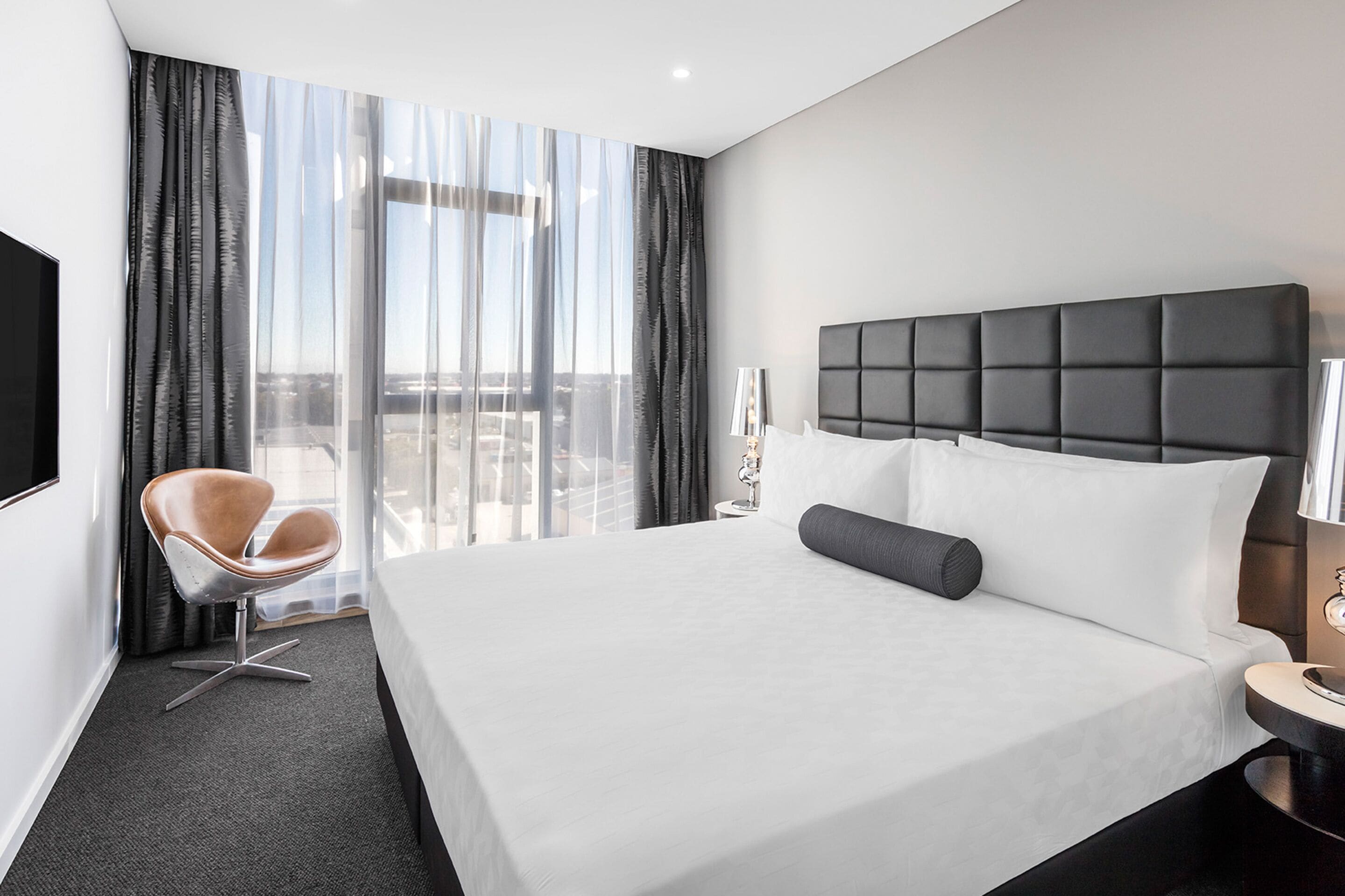 A modern hotel room with a large bed, a chair, a bedside table, and a view of the city through large windows.
