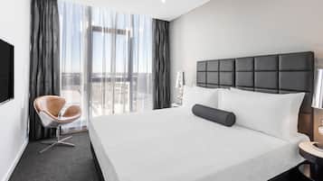 A modern hotel room with a large bed, a chair, a bedside table, and a view of the city through large windows.