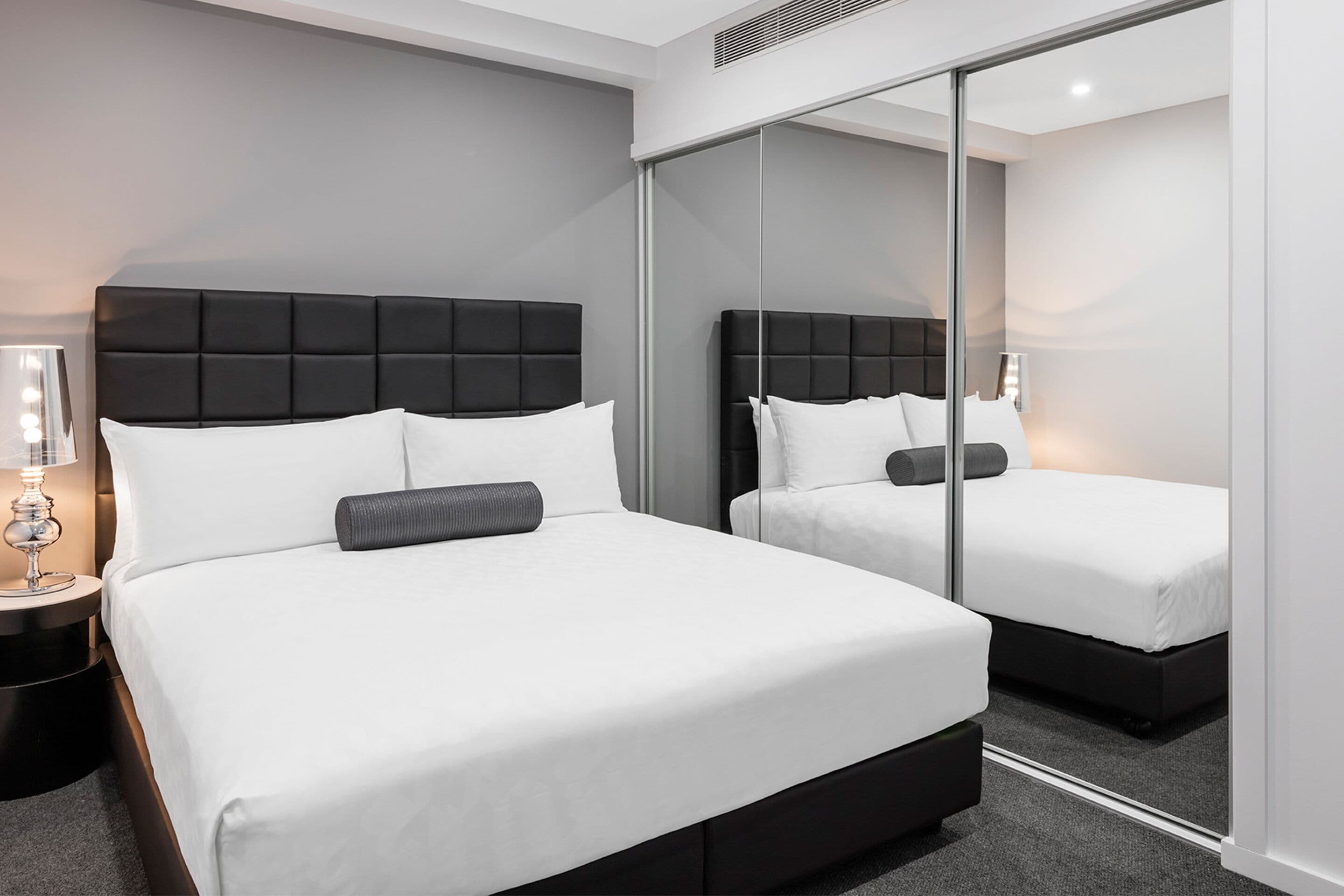 A modern hotel room with a large bed, a nightstand with a lamp, and a mirror wall.