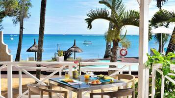 Un área de comedor junto a la playa, con una mesa preparada para dos, con vista al océano y barcos a lo lejos.