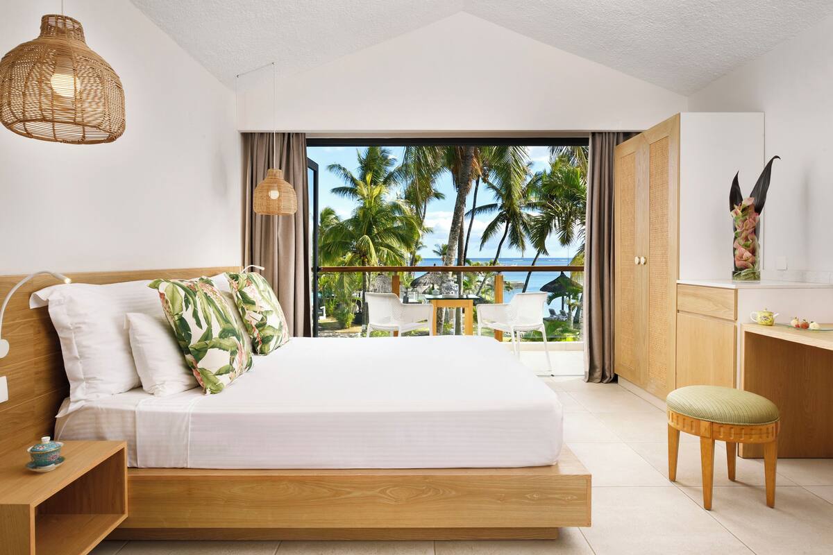 Un dormitorio con una cama grande, cabecera de madera, una mesita de noche, una lámpara colgante y vistas a las palmeras y al océano a través de un ventanal amplio.