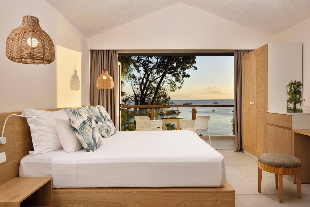 Un dormitorio con una cama grande, una mesita de noche de madera, una lámpara colgante de mimbre y vistas al mar a través de una puerta abierta.