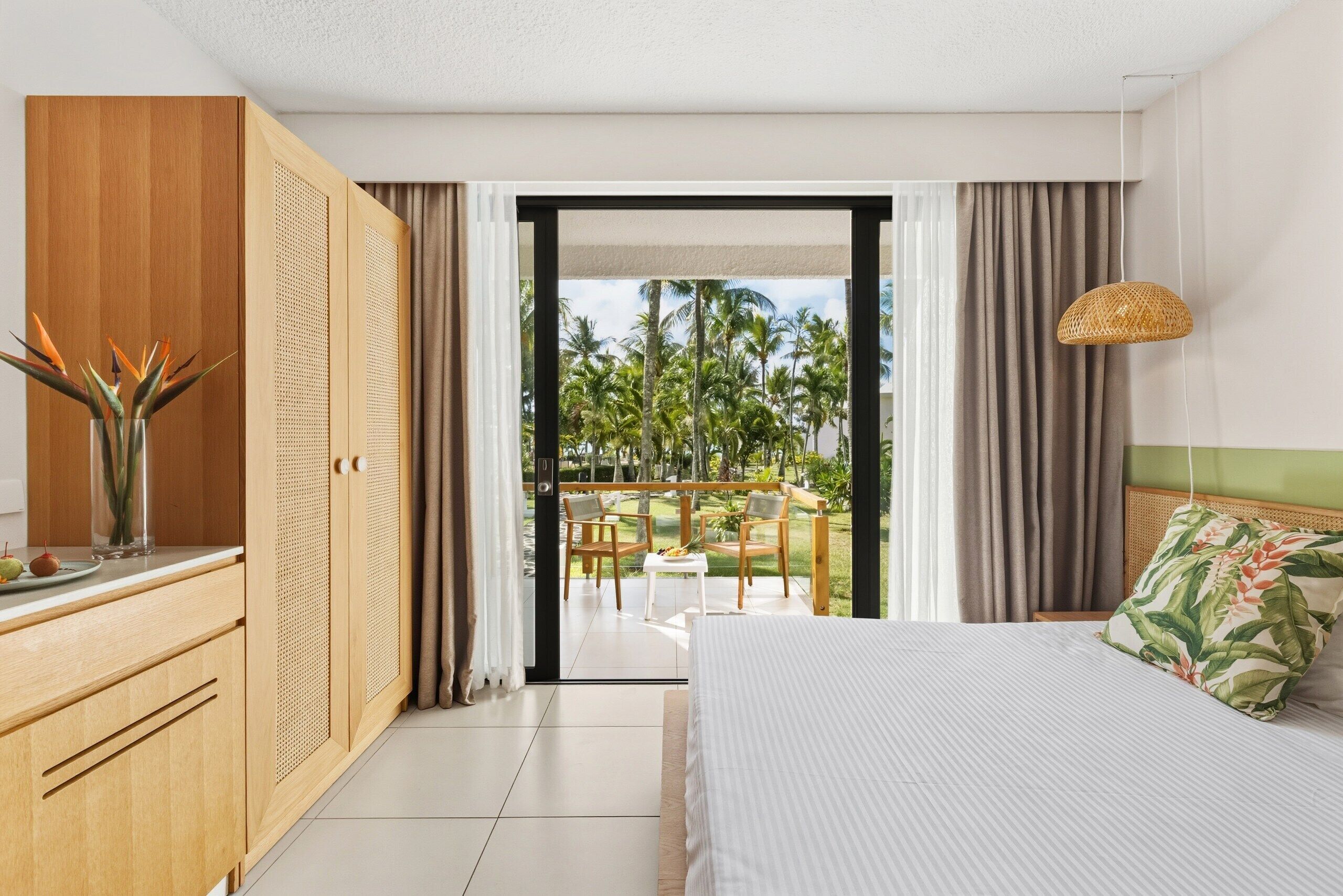 Habitación de hotel con cama, armario de madera, una mesita con un florero y vistas a palmeras a través de una puerta abierta.