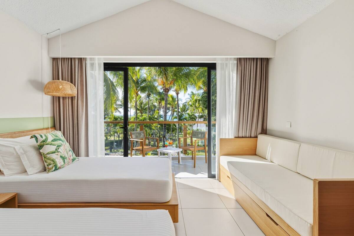 Habitación de hotel con cama, sofá, balcón y vistas a palmeras, además de una lámpara colgante.