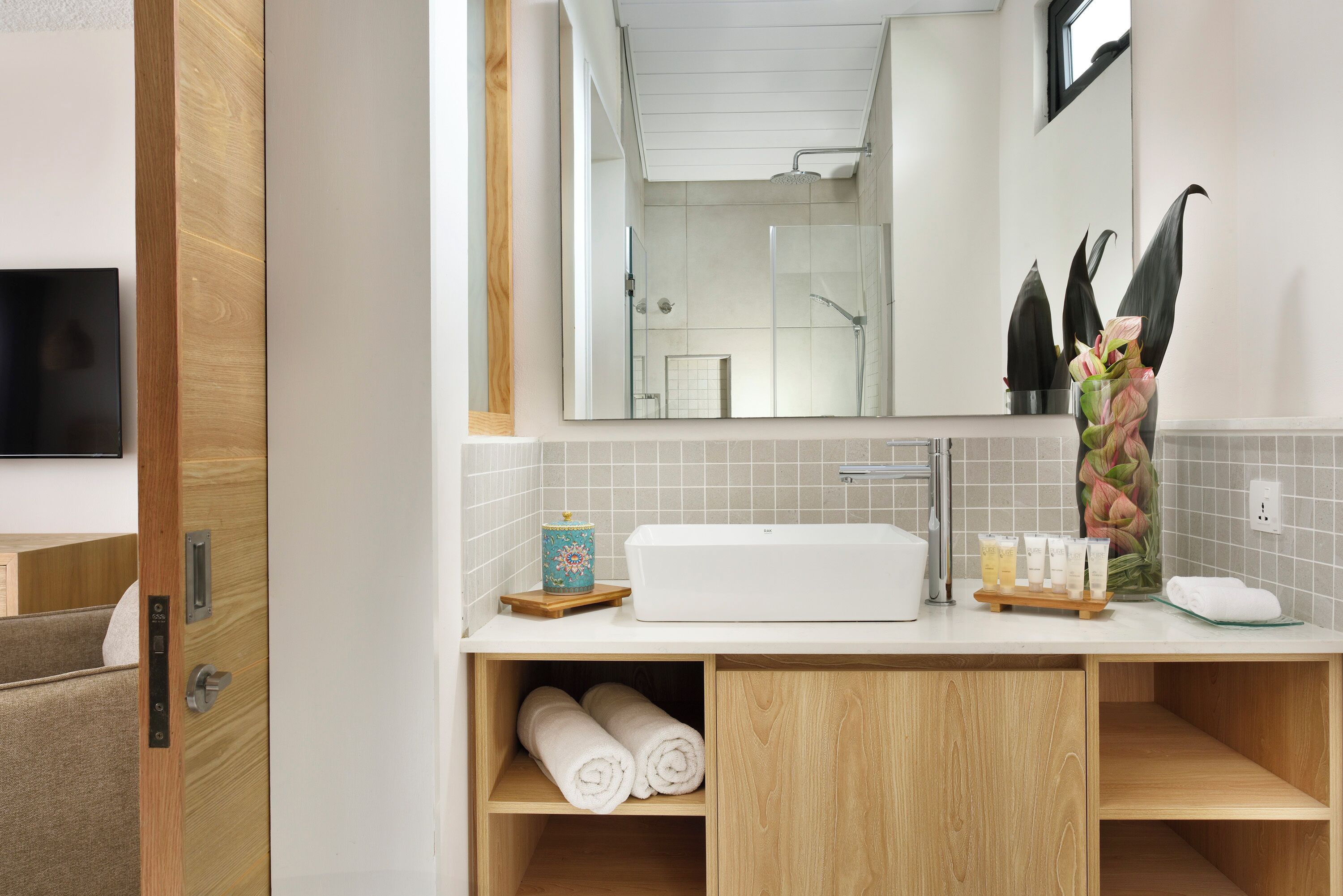 Un baño moderno con un espejo grande, un lavamanos y una ducha. Hay toallas y artículos de aseo en la encimera.
