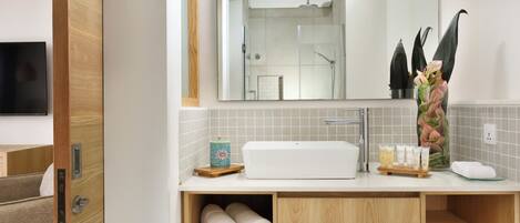 Un baño moderno con un espejo grande, un lavamanos y una ducha. Hay toallas y artículos de aseo en la encimera.