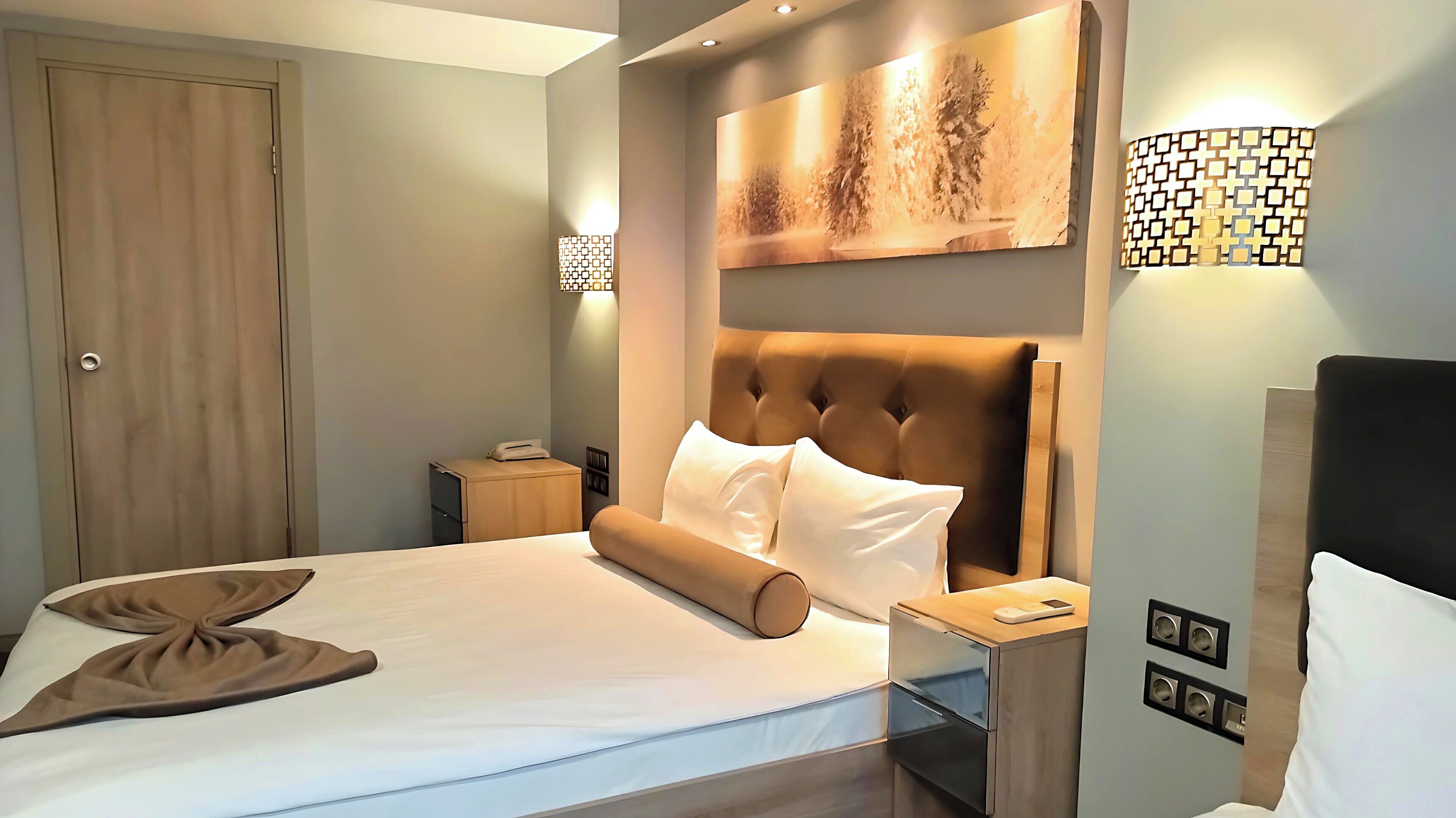 Une chambre d’hôtel avec un grand lit, une tête de lit, une table de chevet, une commode, une lampe fixée au mur et une porte.