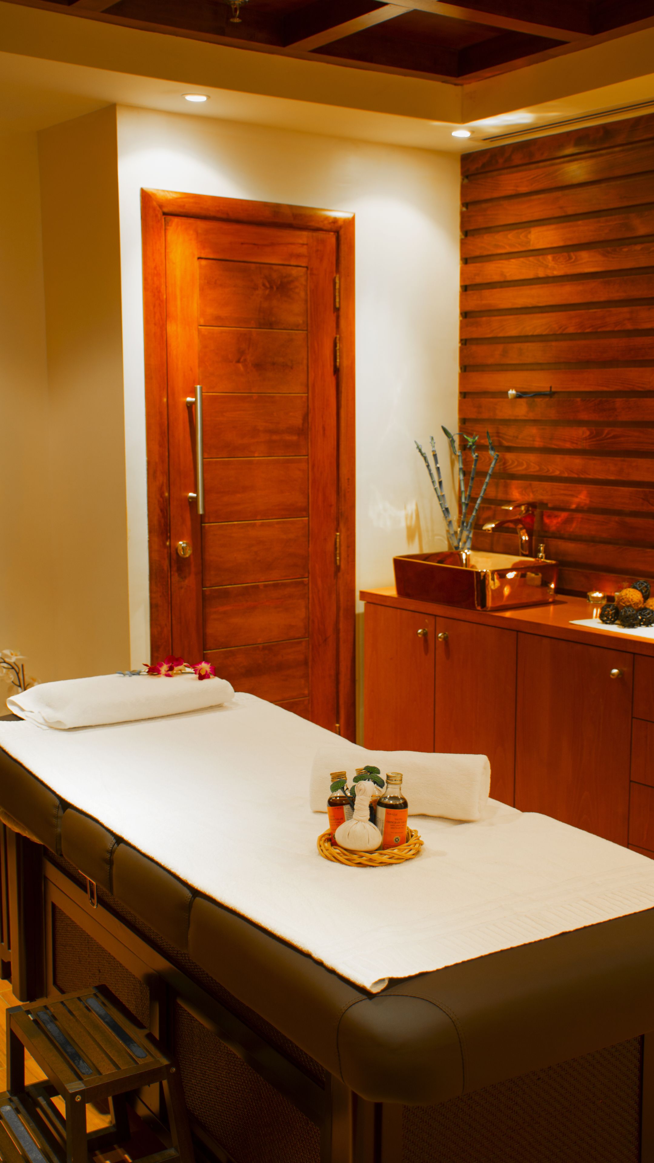 A spa room with a wooden door, a massage table, and a wooden cabinet with bottles and a bowl.