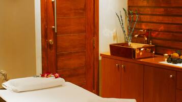 A spa room with a wooden door, a massage table, and a wooden cabinet with bottles and a bowl.
