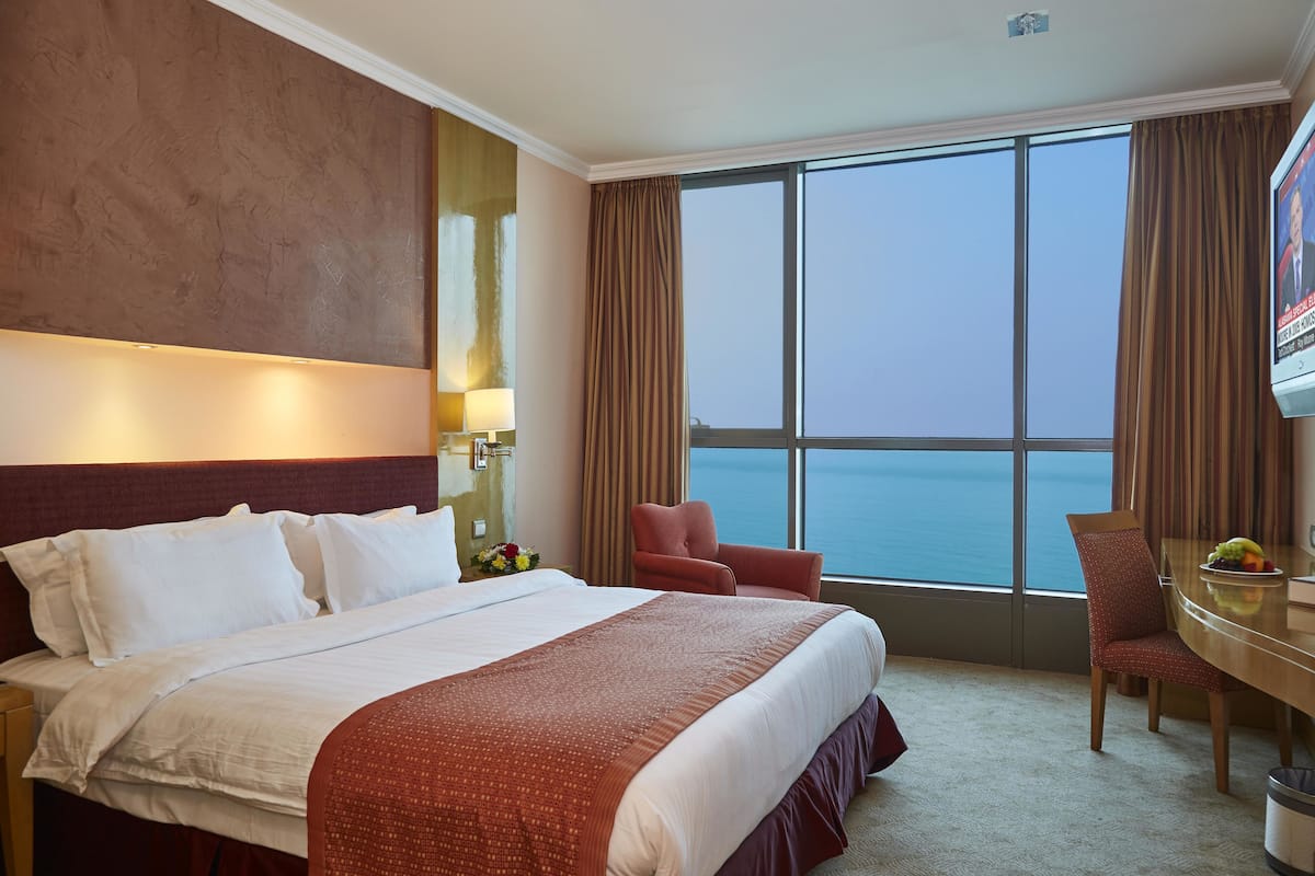 A hotel room with a large bed, a desk with a fruit bowl, a chair, and a view of the sea.