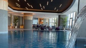 An indoor swimming pool with a waterfall feature, lounge chairs, and a modern interior design. - Grand Hotel (Salmiya)