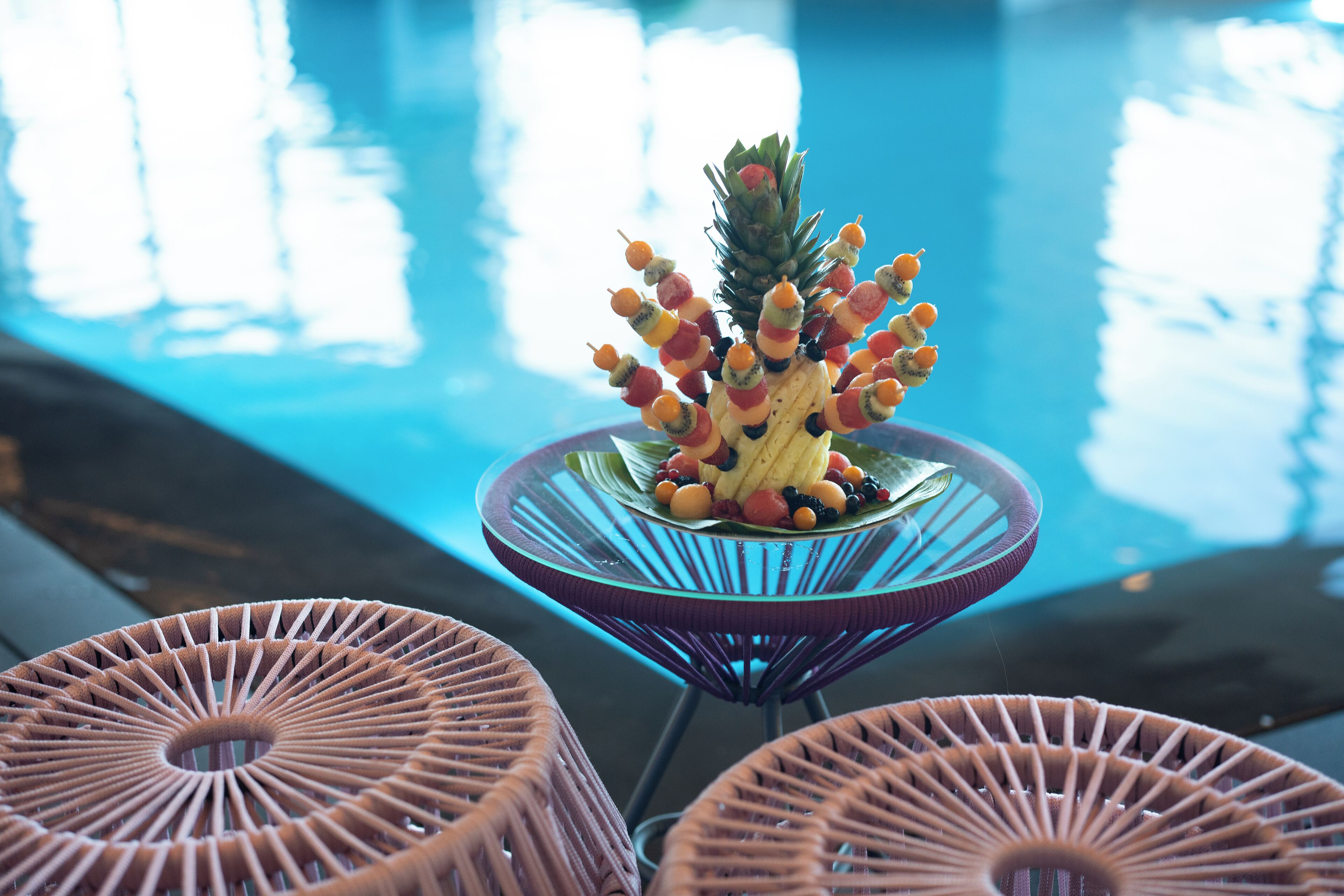 A poolside table with a pineapple centerpiece and fruit skewers.