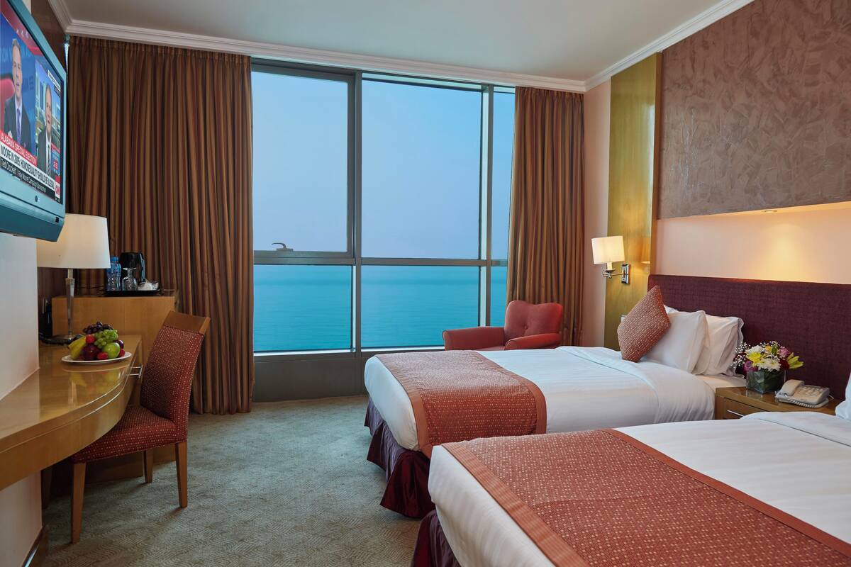 A hotel room with two beds, a desk with a chair, a TV, and a view of the sea.