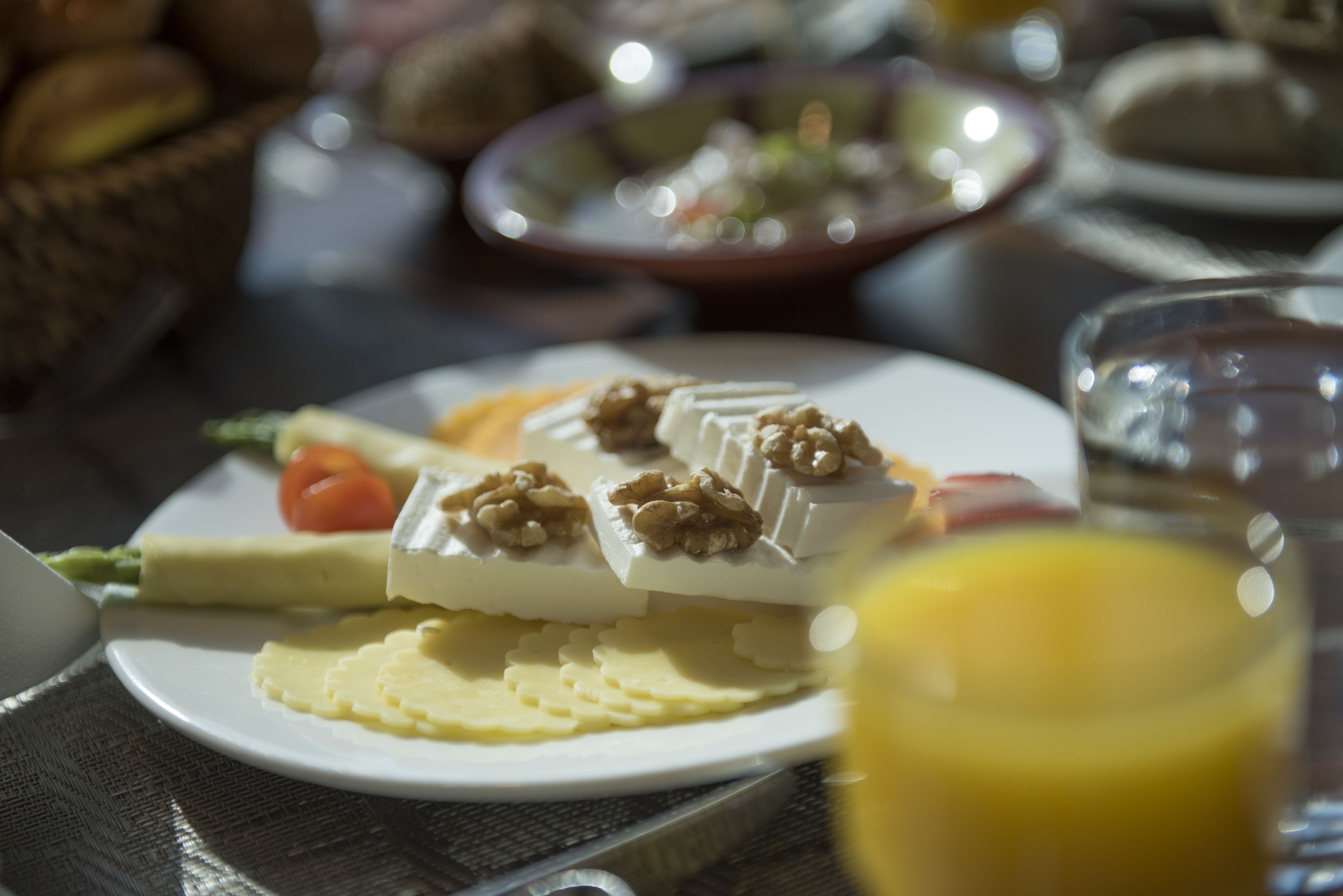 Un desayuno con un plato de queso, tomates y nueces, un vaso de zumo de naranja y una cesta de pan al fondo.