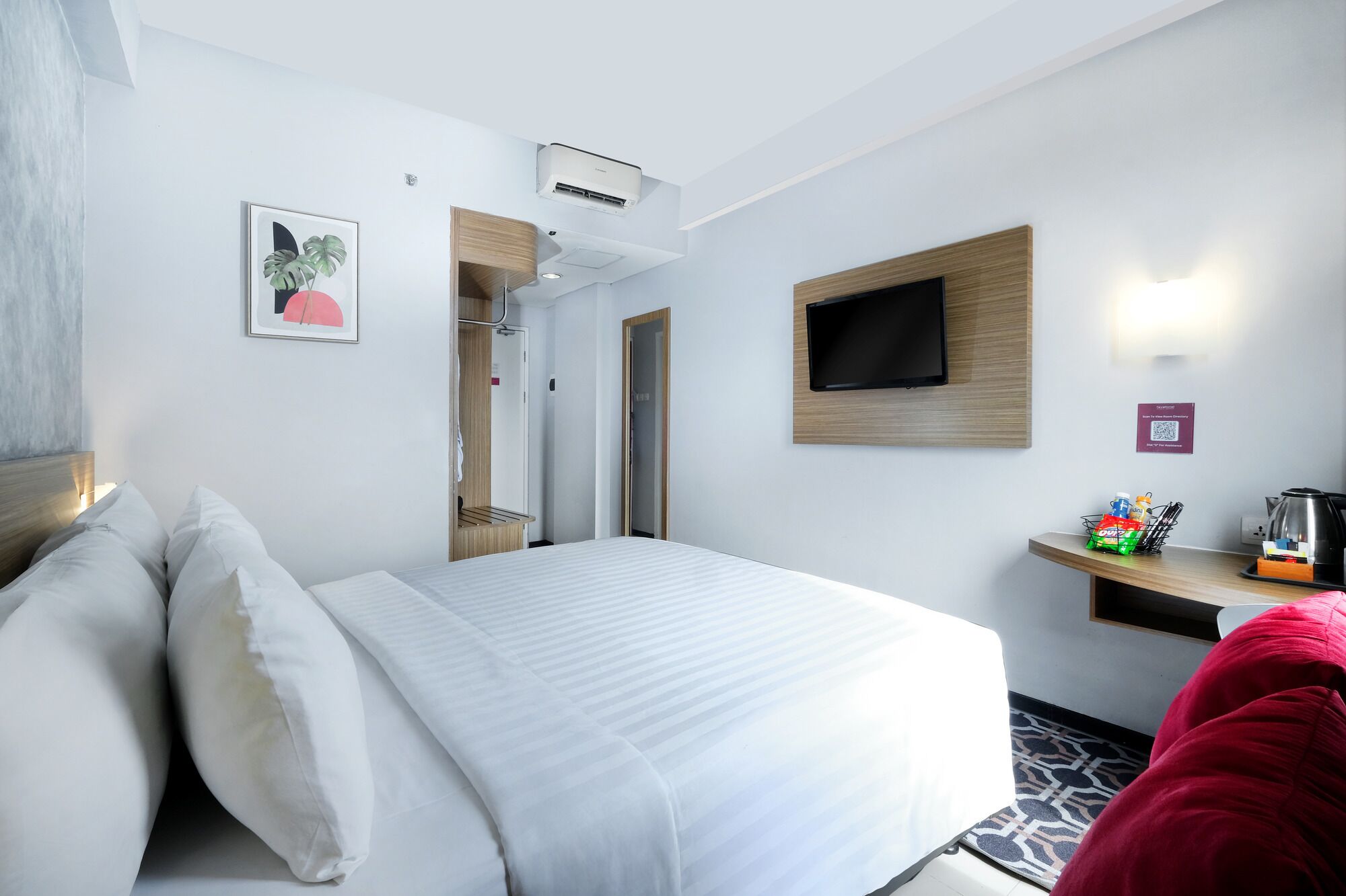 Quarto de hotel com cama, televisão na parede, secretária de madeira e cabideiro.