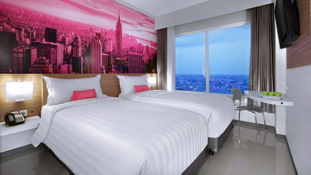 Une chambre d’hôtel avec deux lits, une fresque représentant un paysage urbain, une grande fenêtre donnant sur la ville, un bureau avec une chaise et un téléviseur à écran plat.