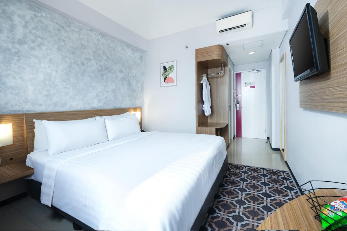 Hotellihuone, jossa on sänky, seinälle asennettu televisio, ilmastointilaite, pyyhekoukku ja kuvioitu lattia.