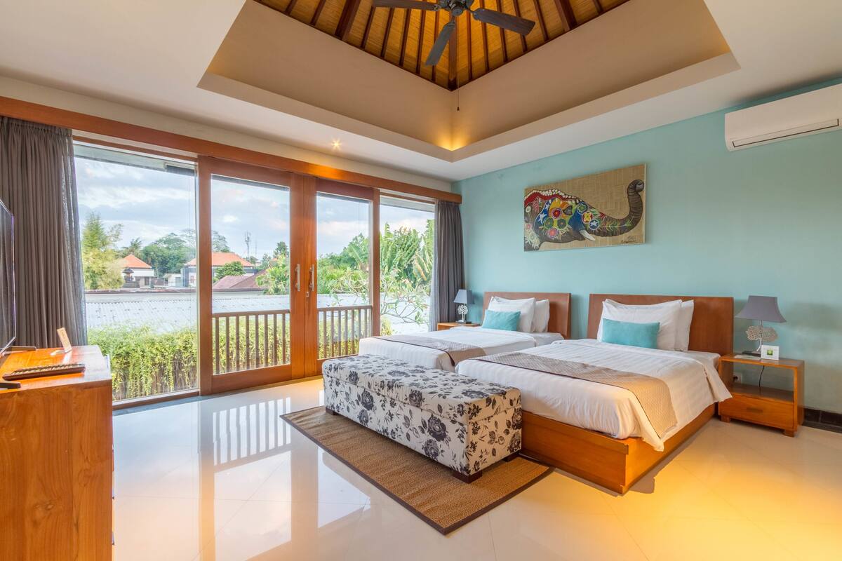 A spacious bedroom with a large bed, a wooden desk, and a balcony with a view of the surrounding area.