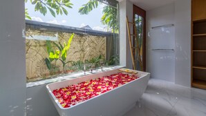 Un baño moderno con una bañera grande llena de pétalos de rosa, una pared de piedra y vistas al exterior con vegetación.