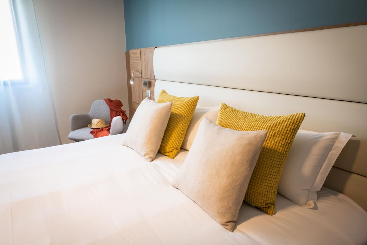 Quarto de hotel com cama, almofadas, uma cadeira e mesa de cabeceira.
