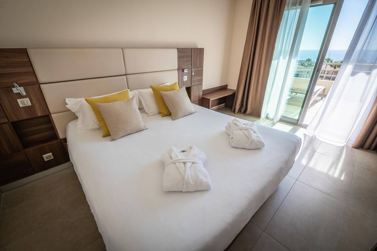 Quarto de hotel com uma cama grande, duas almofadas, uma mesinha de cabeceira e vista para o mar.