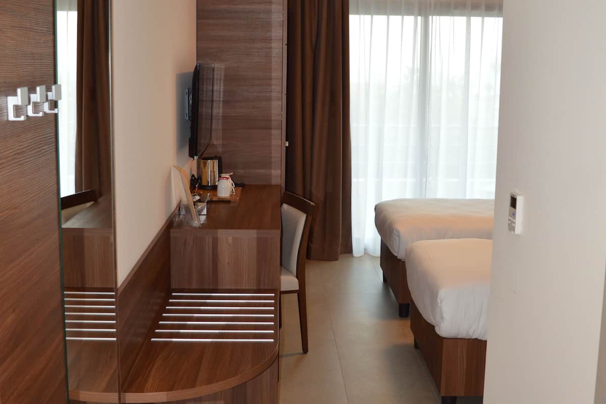 Quarto de hotel com escrivaninha de madeira, uma cama, televisão e uma janela grande com cortinas.