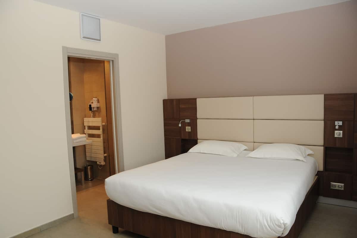 Quarto de hotel com uma cama grande, cabeceira de madeira, mesa de cabeceira e banheiro visível através de uma porta aberta.