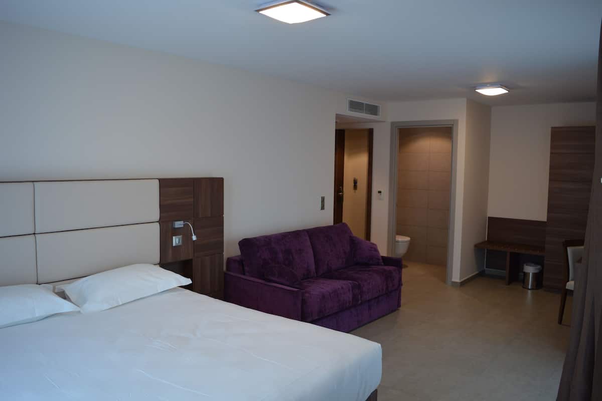 Quarto de hotel com uma cama grande, um sofá roxo, uma mesinha e uma porta que dá para um banheiro.