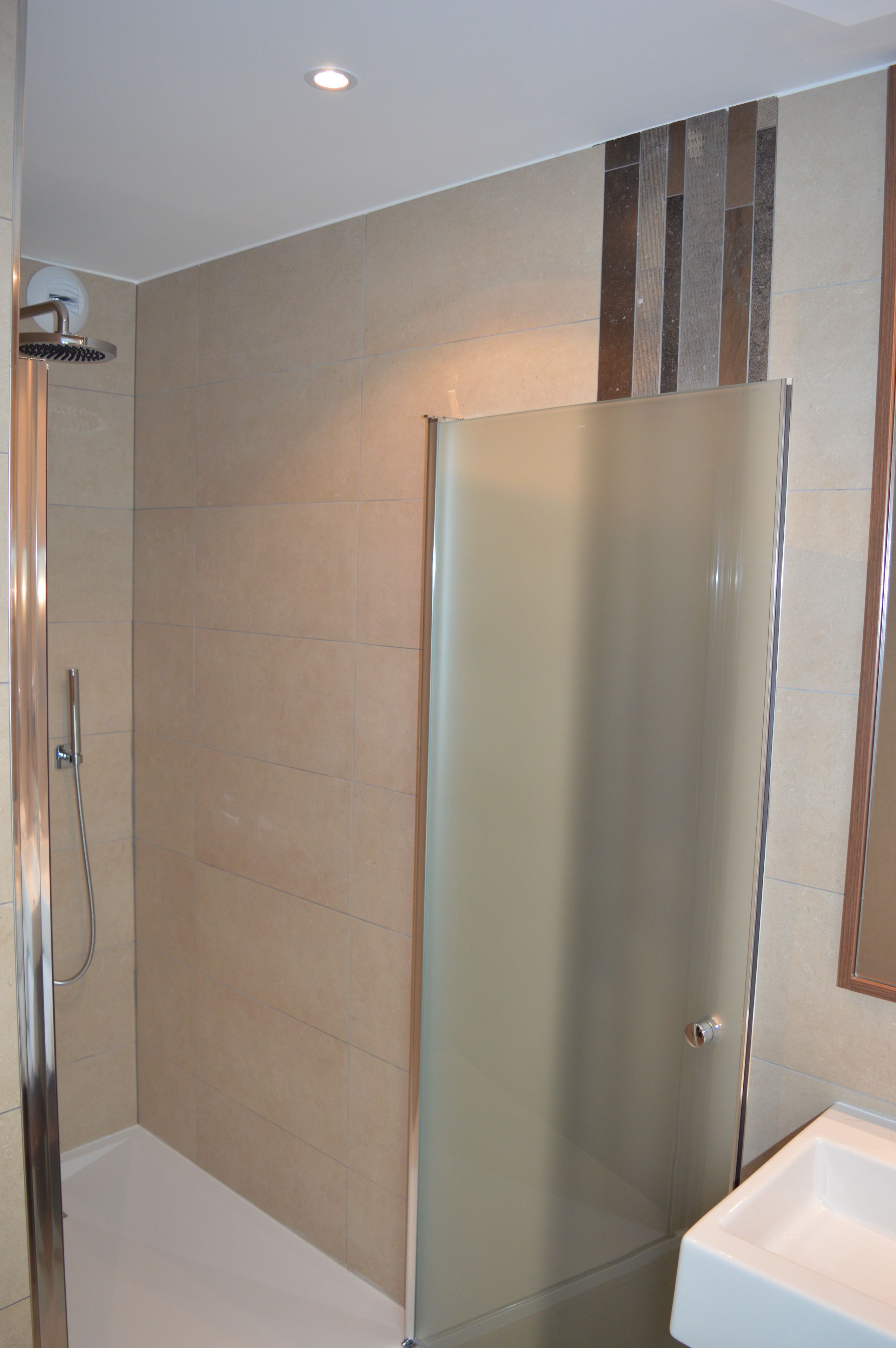 Un baño moderno con ducha envuelta en vidrio, lavamanos blanco y baldosas color beige.