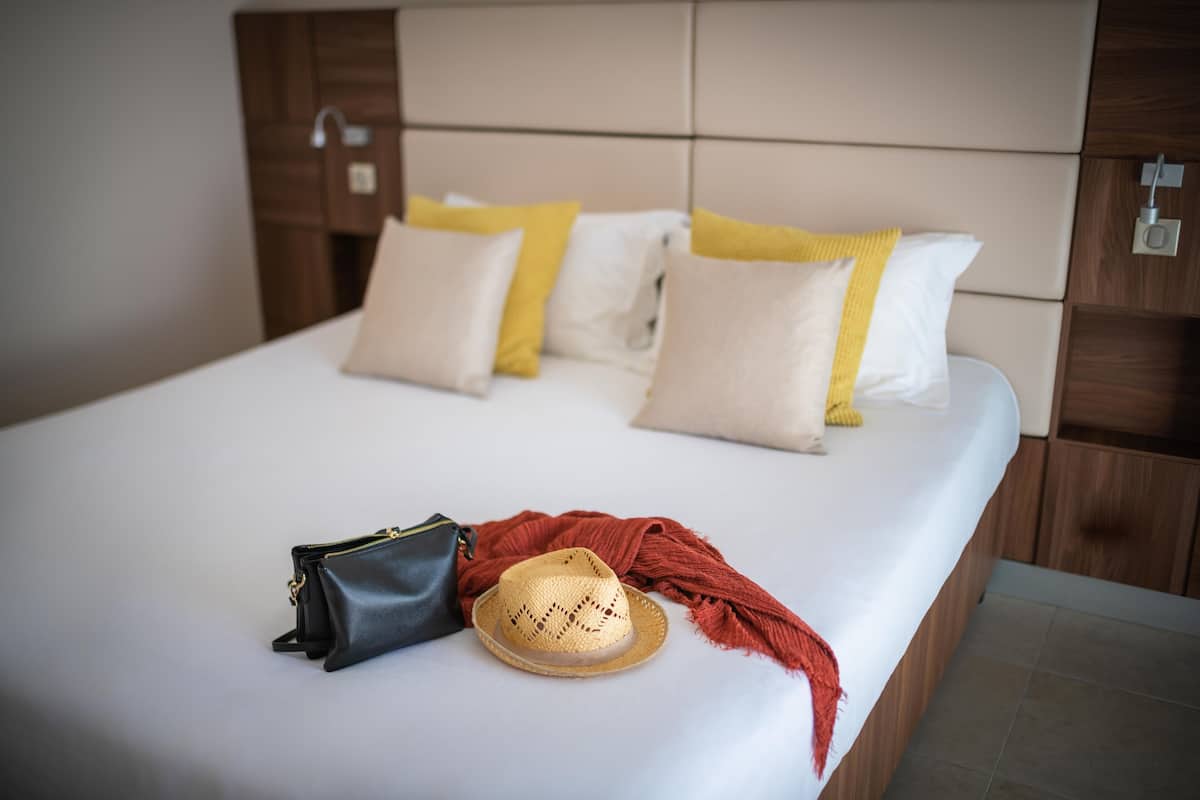 Cama bem arrumada com roupa de cama branca, um chapéu de palha e uma bolsa de mão preta sobre uma superfície limpa.