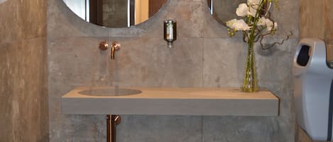 Une salle de bain avec un miroir rond, un lavabo, un vase avec des fleurs et un distributeur de gel hydroalcoolique.