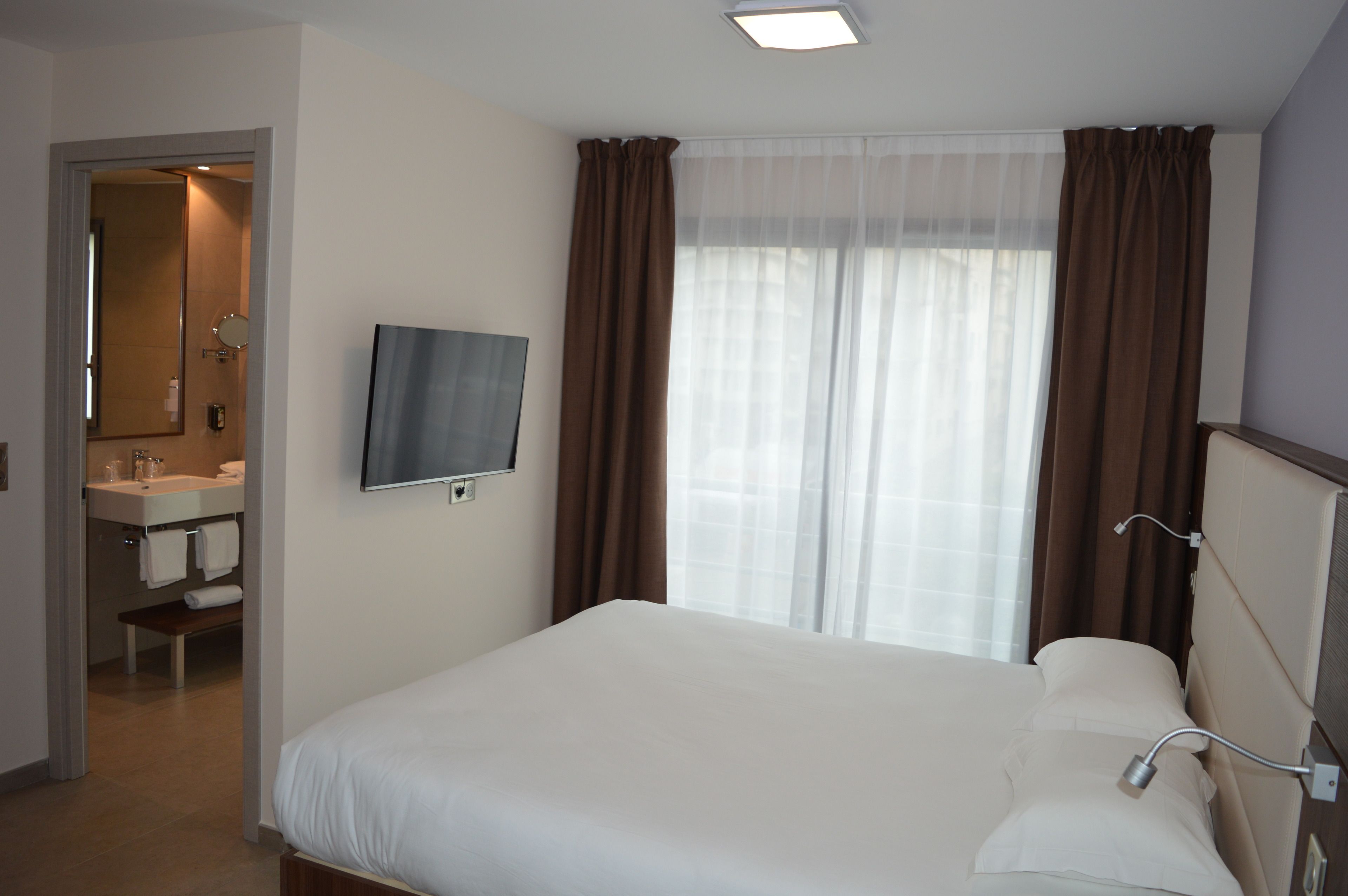 Habitación de hotel con cama, televisión, baño con lavamanos y espejo, y ventana con cortinas.