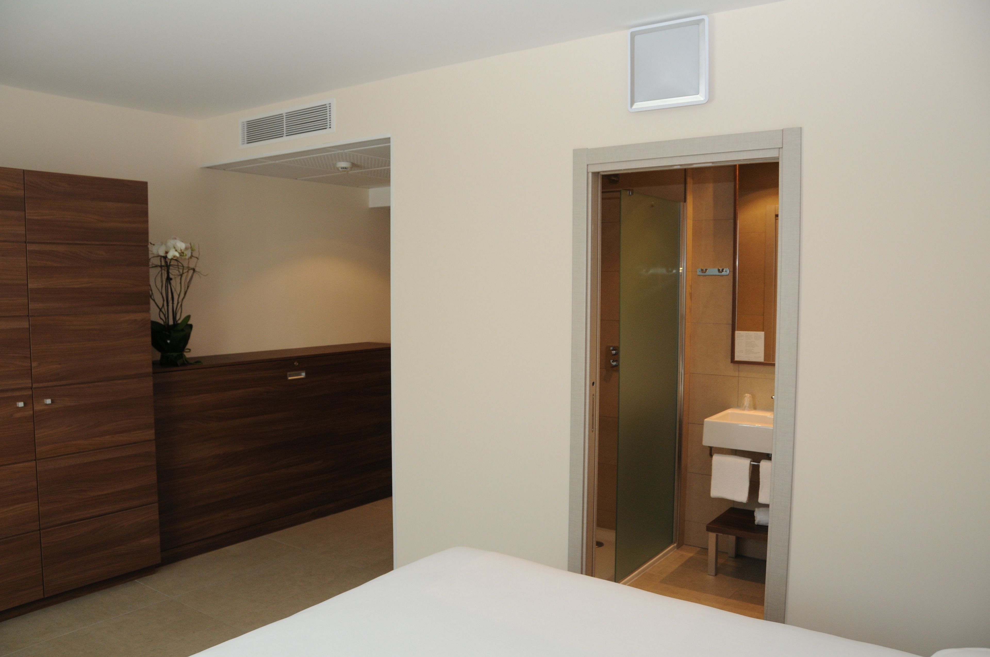 A hotel room with a wooden wardrobe, a small bedside table, a white bed, and a bathroom with a sink and mirror.