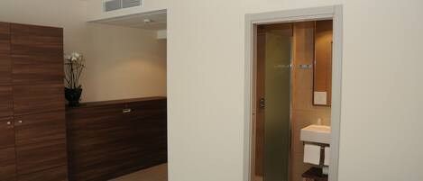 A hotel room with a wooden wardrobe, a small bedside table, a white bed, and a bathroom with a sink and mirror.