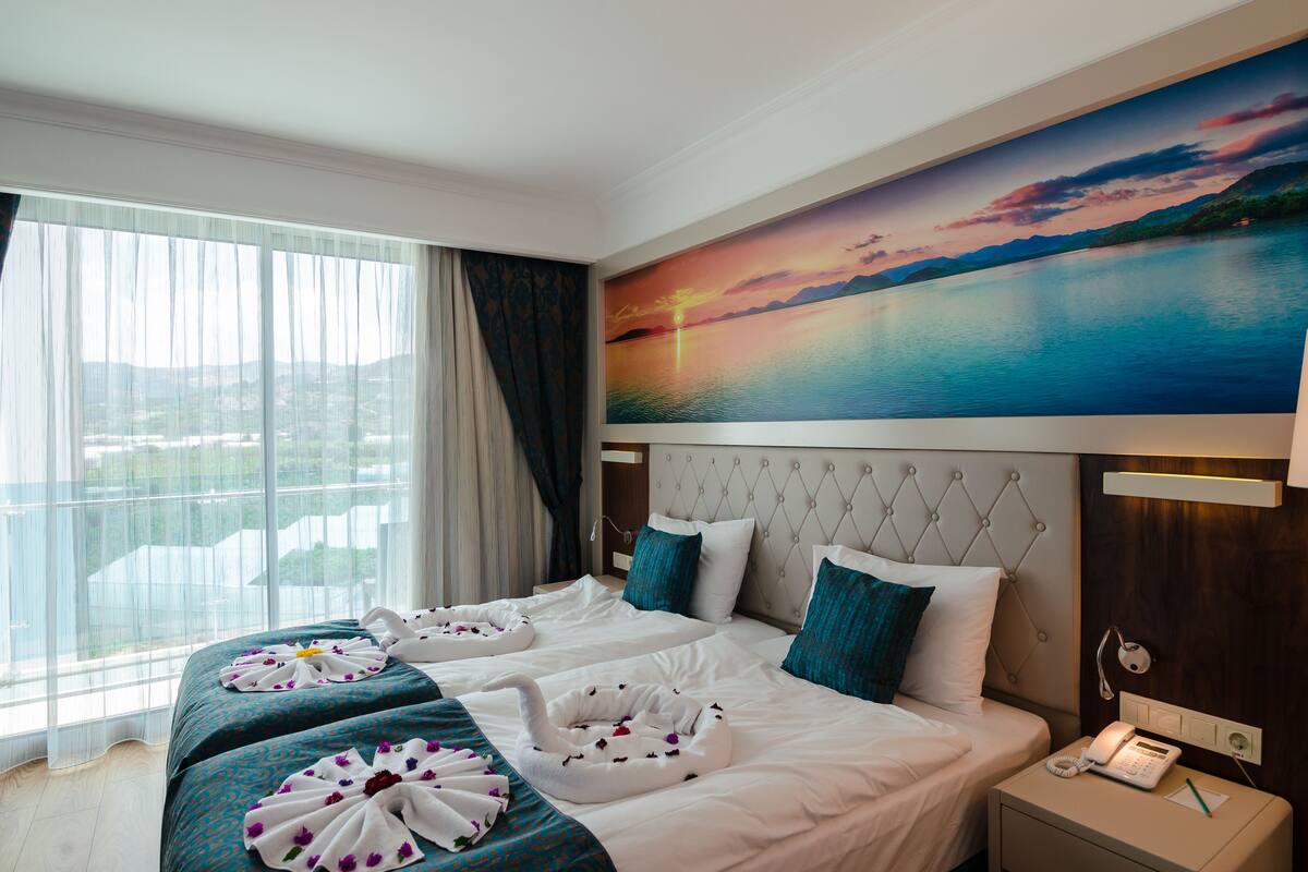A hotel room with a large bed, a painting of a sunset over water, and a view of the city through the window.