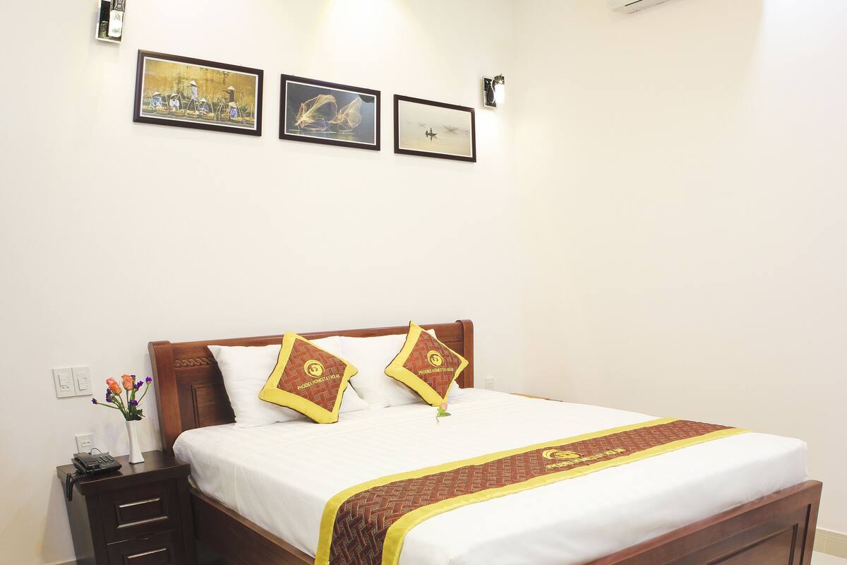 A neatly made bed with a wooden frame, white bedding, and decorative pillows. There is a bedside table with a phone and a vase of flowers. Three framed artworks are hung on the wall above the bed.