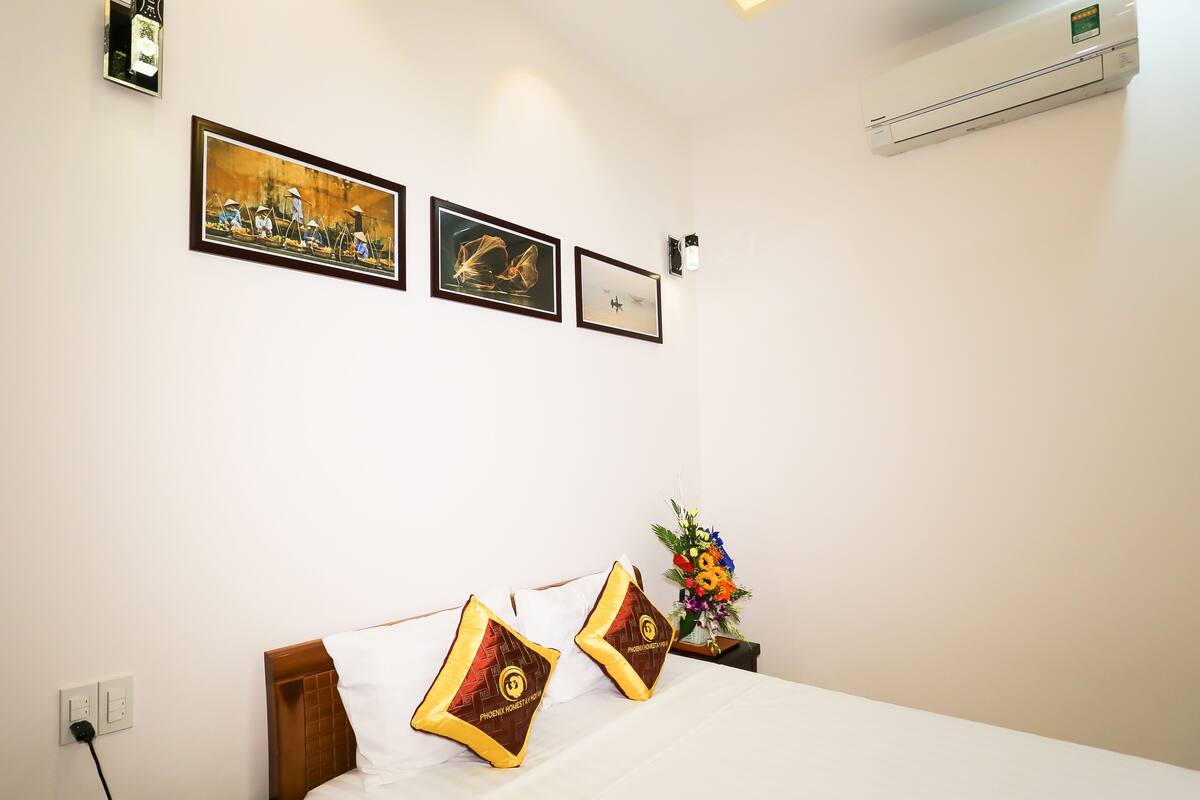 A hotel room with a bed, two pillows, a nightstand with a vase of flowers, and three framed pictures on the wall.