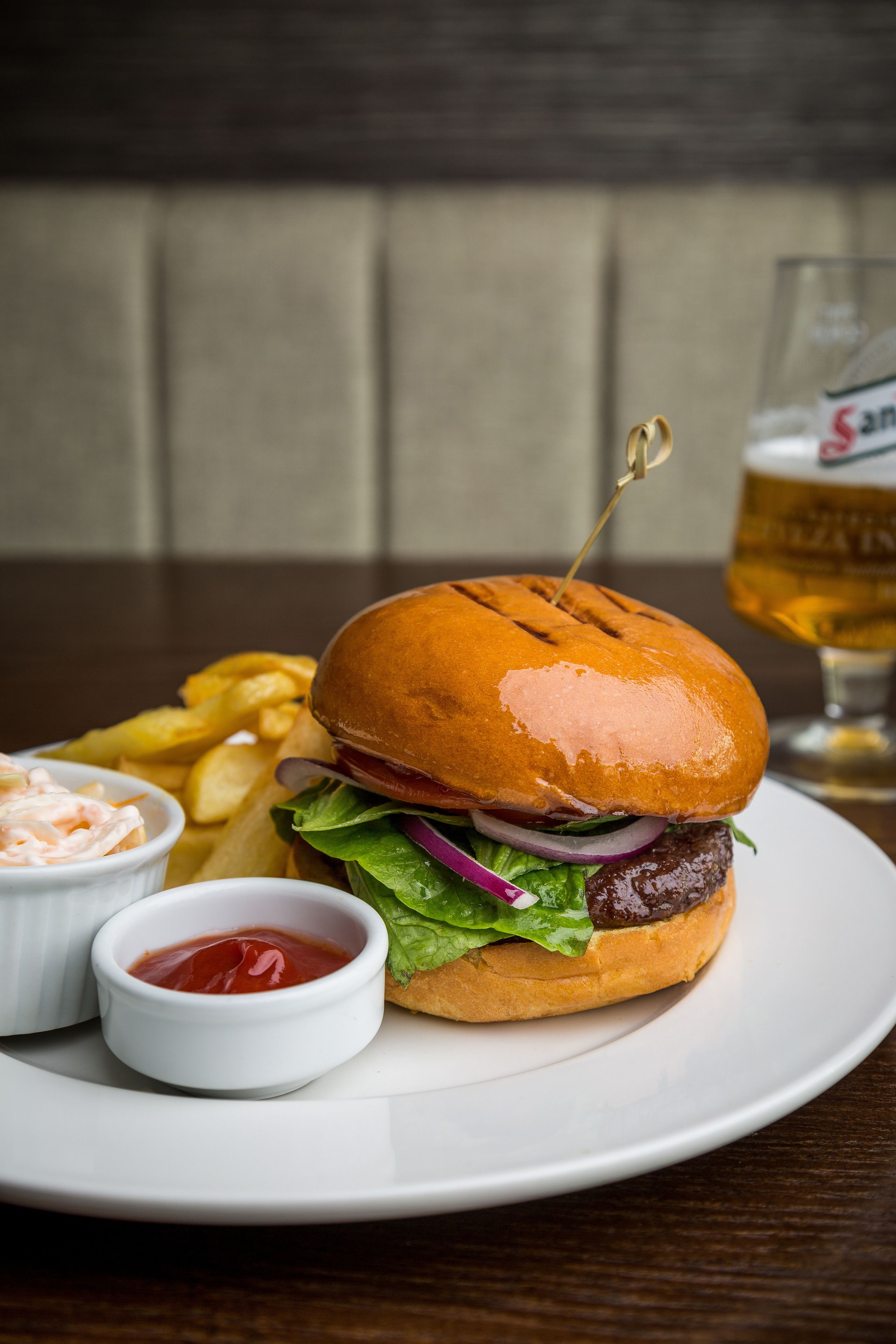 Hamburguesa con papas fritas y kétchup en un plato, con un vaso de cerveza al fondo.