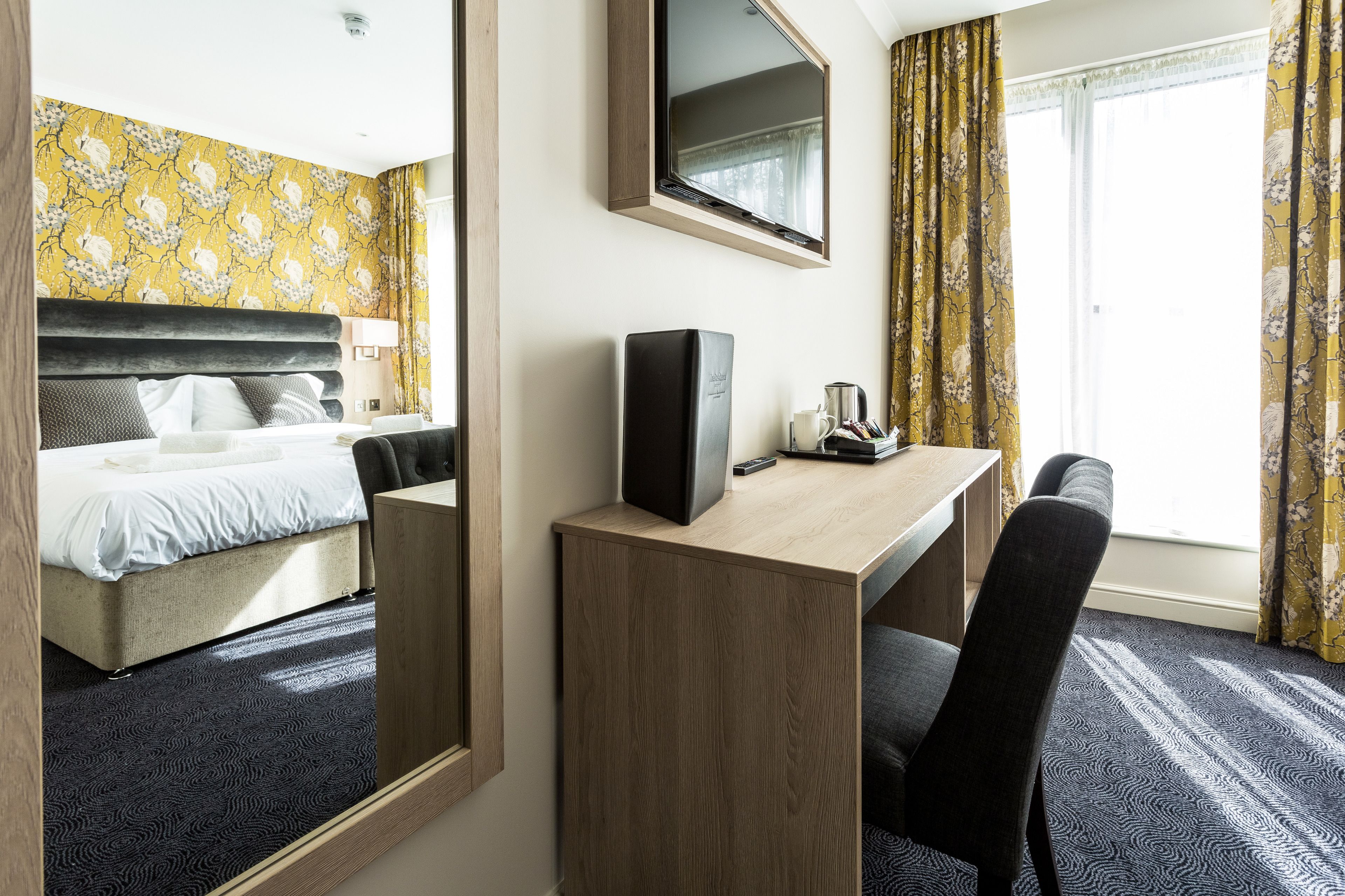 Habitación de hotel con cama, escritorio con computadora, silla, espejo y ventana con cortinas.