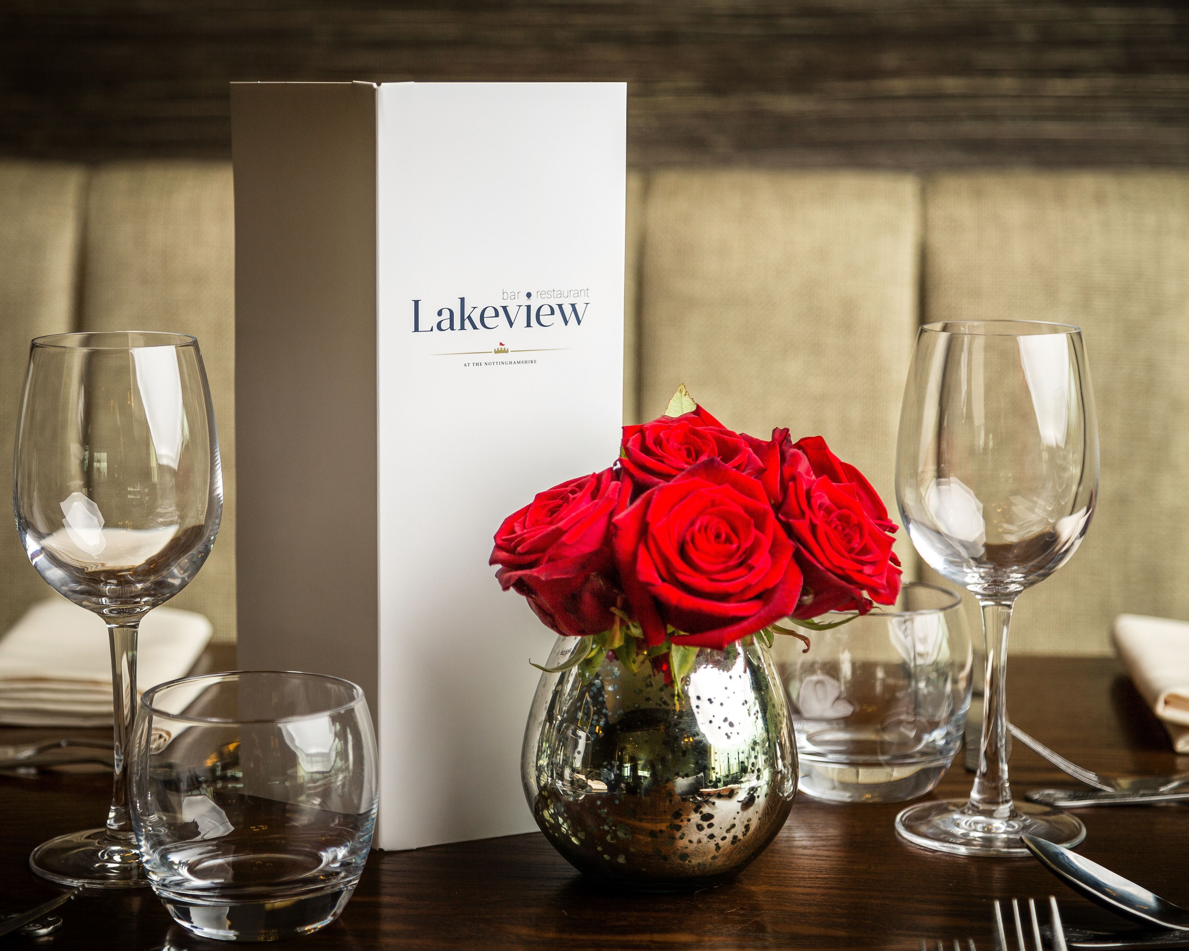 A table setting with a menu box labeled "Lakeview bar restaurant" and a vase with red roses.